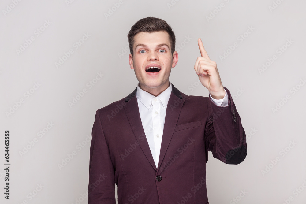 Portrait of handsome young man raises fore finger as gets idea or remembers to do something not in time, wearing violet suit and white shirt. Indoor studio shot isolated on grey background. Portrait of handsome young man raises fore finger as gets idea or remembers to do something not in time, wearing violet suit and white shirt. Indoor studio shot isolated on grey background.