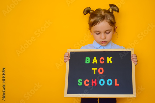 Child holding black board with colored letters saying back to school, on orange background. Little cute schoolgirl smiling and wanting to learn. Waiting for the school year to start