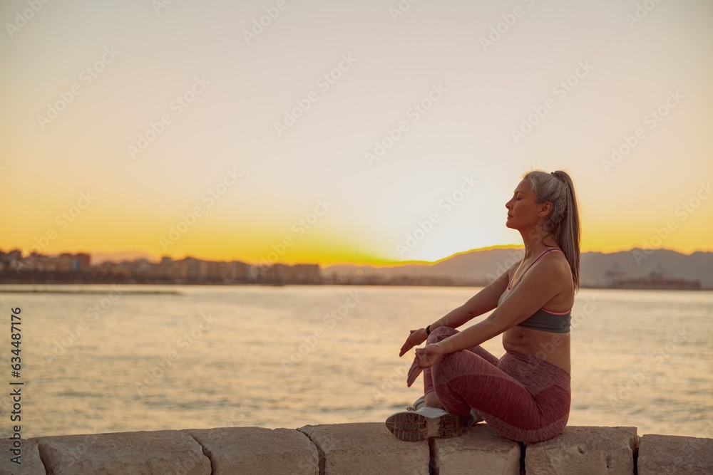 Beautiful mature woman sitting by the sea in lotus pose, meditating with clsed eyes on background with sunset and mountains