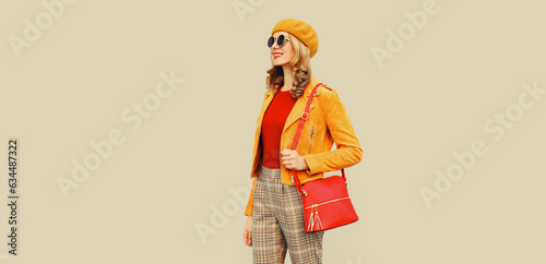 Autumn style outfit, beautiful stylish young woman with handbag looking away wearing orange french beret hat, jacket and round sunglasses on gray background