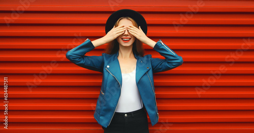 Portrait of stylish happy surprised smiling young woman covering her eyes wearing black round hat, blue leather jacket on red background