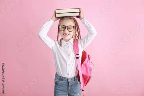 Little smiling girl in glasses with the book on her head. Clever schoolchild with funny hairdo pigtails in white shirt, backpack on pink background. Learning and education of kid