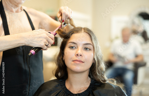 In beauty salon, cropped unrecognizable elderly woman hairdresser makes styling curls for young girl client. Hairdresser uses curling iron to create festive image of young female client