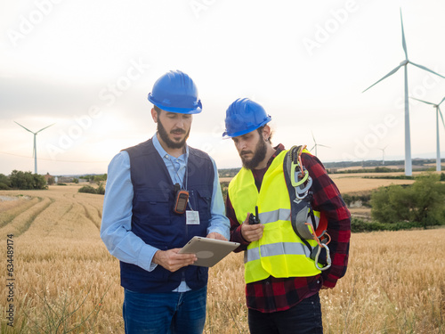 Two male engineers work and share ideas with a tablet in a field of wind turbines