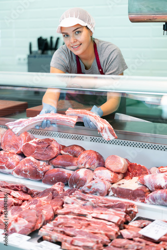 Young woman butcher in apron lays out raw beef ribs on counter in butcher shop