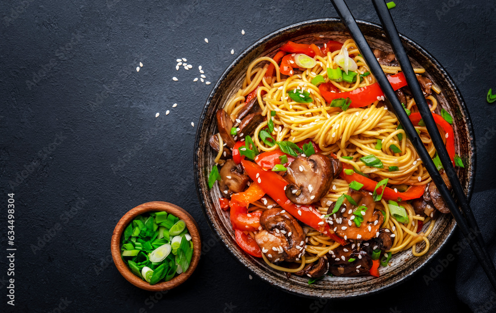 Stir fry noodles with vegetables: red paprika, champignons, green onion and sesame seeds in ceramic bowl. Black table background, top view