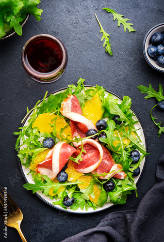 Delicious salad with smoked duck, oranges, blueberries and arugula on black table background, top view