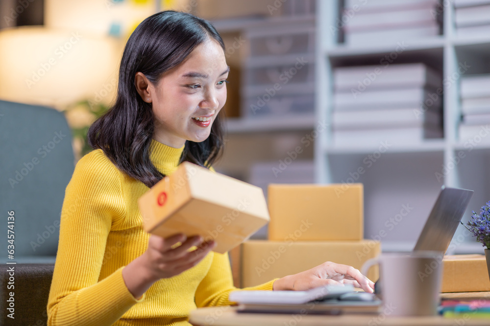 Startup small business entrepreneur or freelance Asian woman using a laptop with box in night, Young success Asian woman with her hand lift up, online marketing packaging box and delivery, SME concept