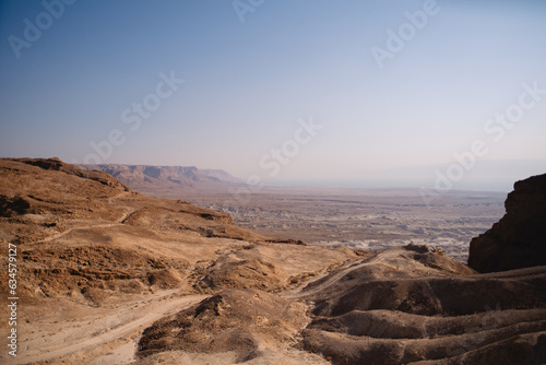 A lonely unpaved road passes over some hills in the Judean Desert, overlooking cliffs and an expansive valley and the Dead Sea. The hazy air adds a sense of depth to the barren landscape.