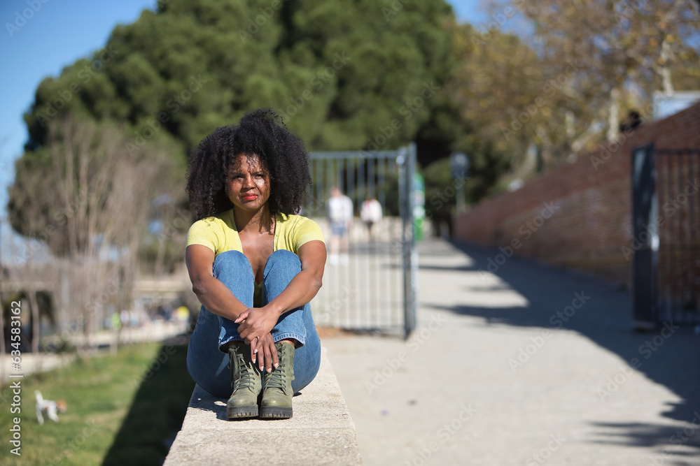 Young, beautiful, black woman with afro hair, wearing yellow t-shirt, jeans and boots, sitting on a stone wall, clutching her knees, calm and relaxed. Concept relax, tranquility, peace.