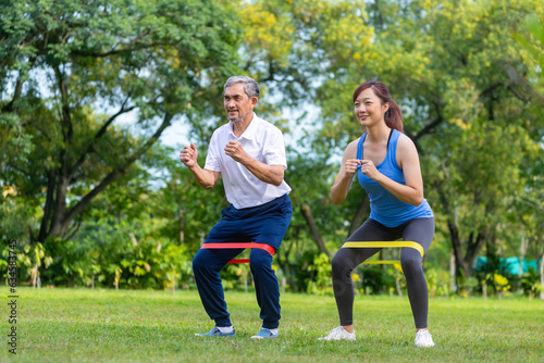 Senior asian man and his daughter are using sport rubber band to build up his leg muscle strength in the public park for elder longevity exercise and outdoor workout