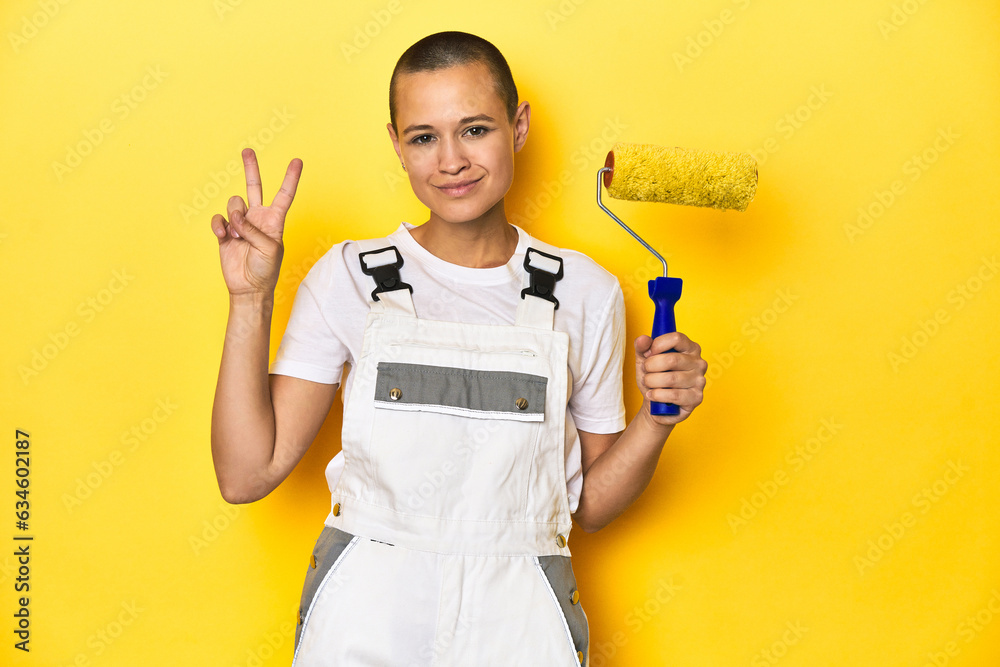 Painter woman, shaved head, with yellow roller in studio showing number two with fingers. Painter woman, shaved head, with yellow roller in studio showing number two with fingers.