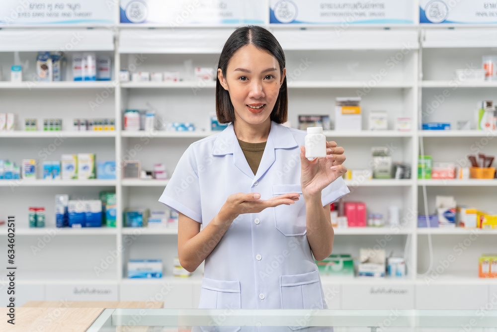 Happy beautiful asian female pharmacist wearing lab coat standing and holding pill bottle, She looking at camera feels good, trustworthy and proud of his work in the pharmacy drugstore.