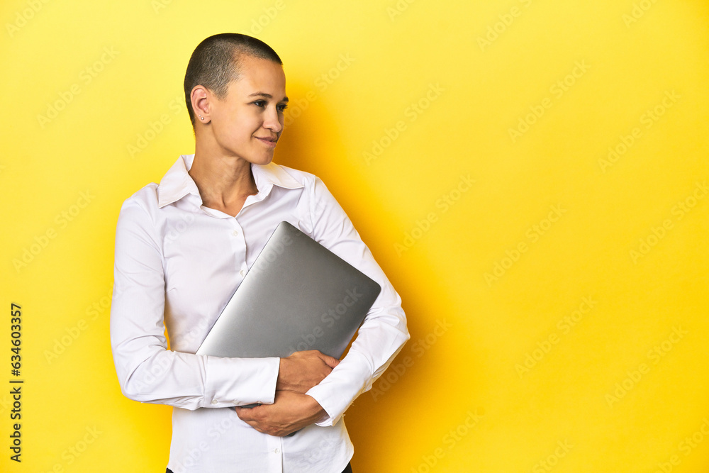 Woman with shaved head holding laptop, yellow studio background Woman with shaved head holding laptop, yellow studio background