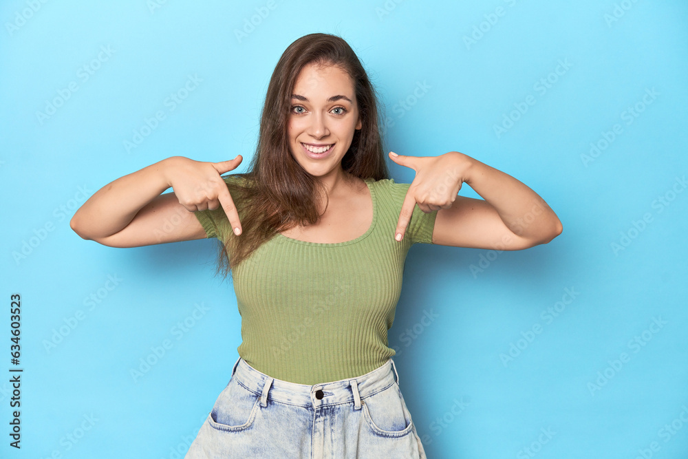 Young Caucasian woman in a green top on a blue backdrop points down with fingers, positive feeling. Young Caucasian woman in a green top on a blue backdrop points down with fingers, positive feeling.