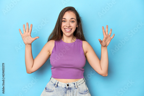 Fashionable young woman in a purple top on blue background showing number ten with hands.