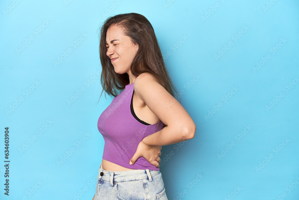 Fashionable young woman in a purple top on blue background suffering a back pain. Fashionable young woman in a purple top on blue background suffering a back pain.