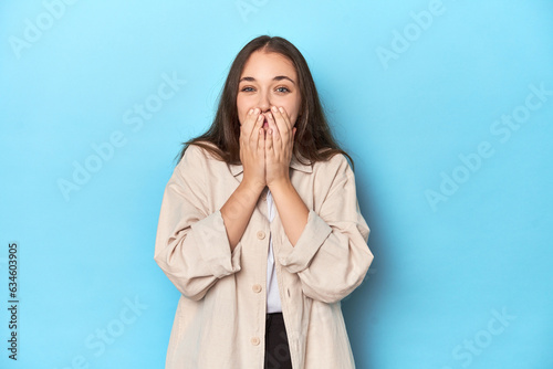 Stylish young woman in an overshirt on a blue background laughing about something, covering mouth with hands.