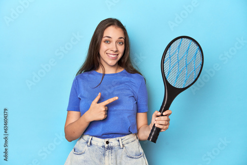 Woman with an electric bug zapper on a blue background smiling and pointing aside, showing something at blank space.