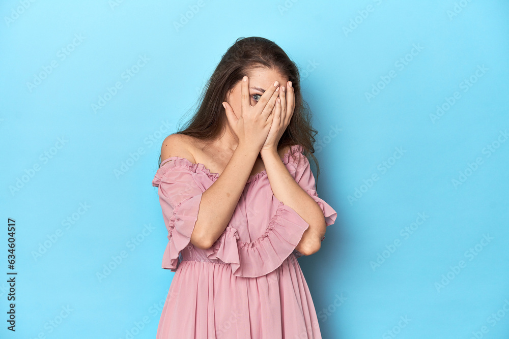 Young woman wearing a pink dress on a blue studio backdrop blink through fingers frightened and nervous. Young woman wearing a pink dress on a blue studio backdrop blink through fingers frightened and nervous.