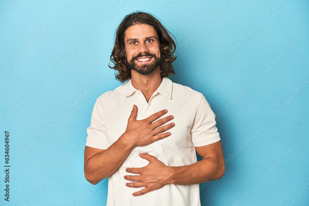 Long-haired man in a white polo, blue studio laughs happily and has fun keeping hands on stomach. Long-haired man in a white polo, blue studio laughs happily and has fun keeping hands on stomach.