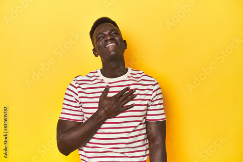 Stylish young African man on vibrant yellow studio background, laughs out loudly keeping hand on chest.