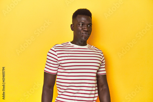 Stylish young African man on vibrant yellow studio background, shrugs shoulders and open eyes confused.