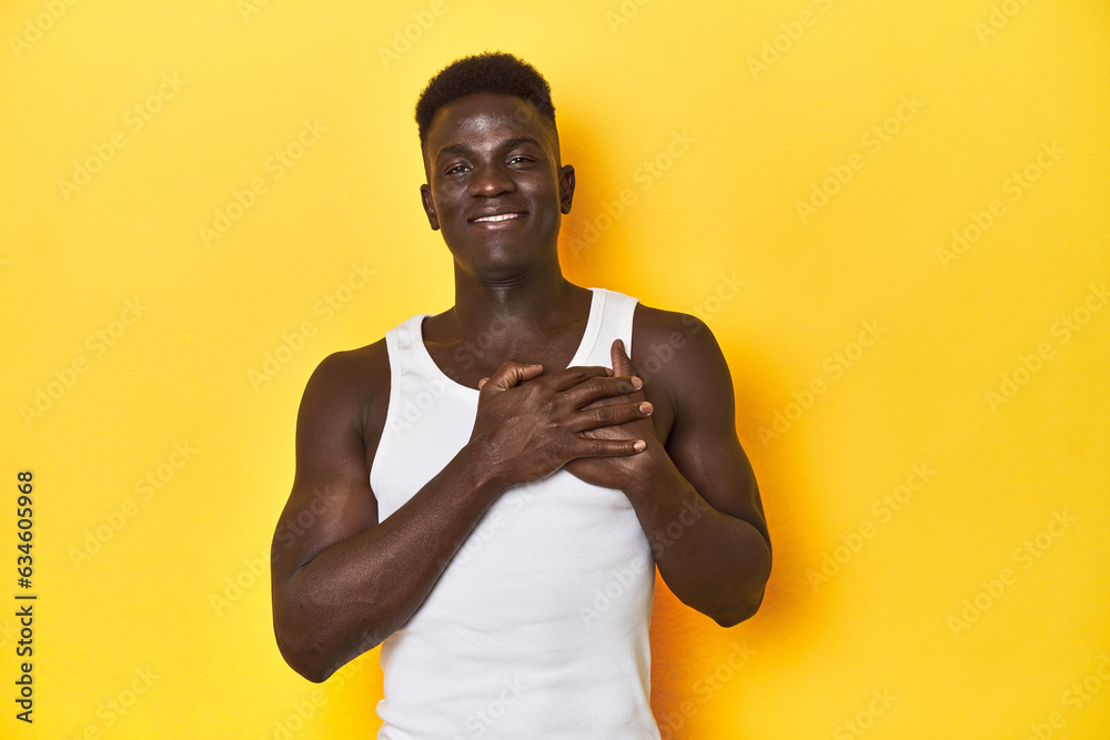 Stylish young African man on vibrant yellow studio background, laughing keeping hands on heart, concept of happiness.