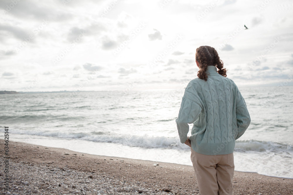 Young beautiful girl walking in the sand beach in cold weather day. Woman dressed warm clothes. Back view of the woman Young beautiful girl walking in the sand beach in cold weather day. Woman dressed warm clothes. Back view of the woman
