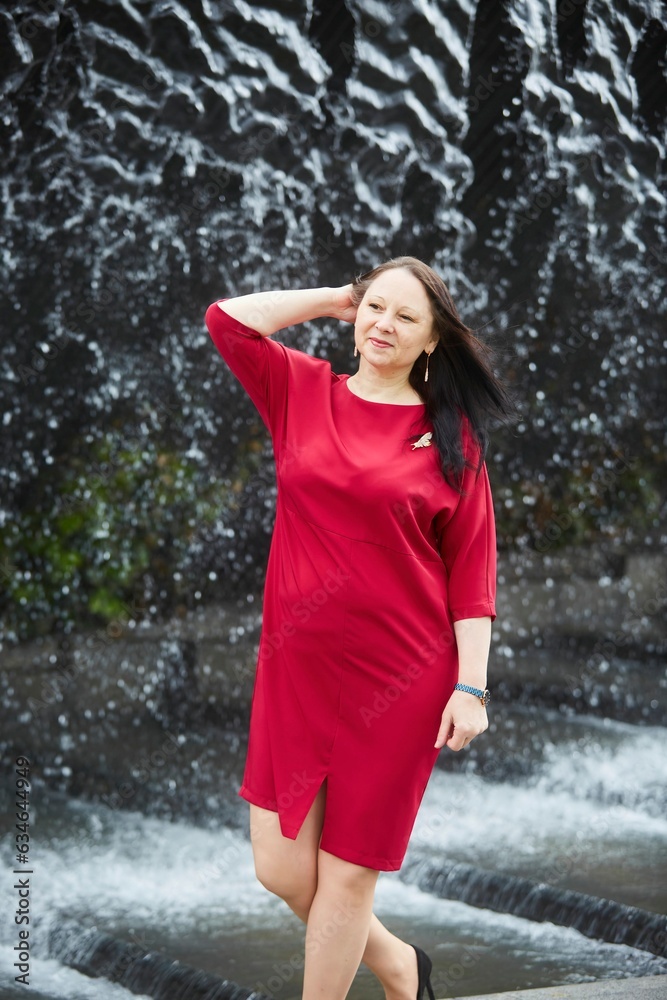 A spectacular older woman in a bright red dress on the background of a waterfall in the park. A spectacular older woman in a bright red dress on the background of a waterfall in the park.