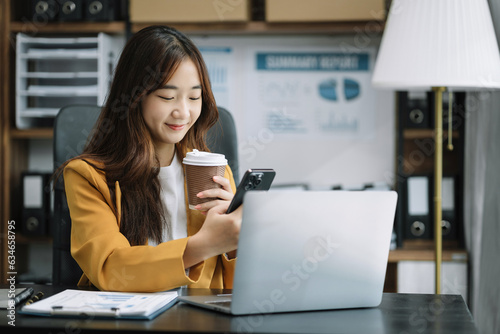 Asian businesswoman sits in a office working on smartphone and enjoys a coffee.