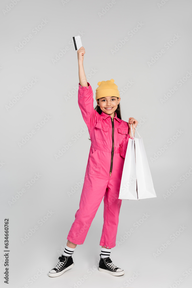 positive girl in beanie hat and trendy outfit holding credit card on grey, kid shopping, fashion positive girl in beanie hat and trendy outfit holding credit card on grey, kid shopping, fashion
