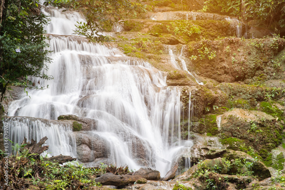 Fototapeta premium Natural waterfall view flowing through the rocky mountain with tropical forest and moss in sun light effect and dark green tone