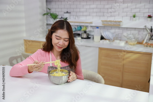 Portrait of young happy asian currly hair woman sitting on table and eating tasty and instant noodle for lunch in the kitchen with chopsticks and brick wall. Unhealthy and cheap food lifestyle concept