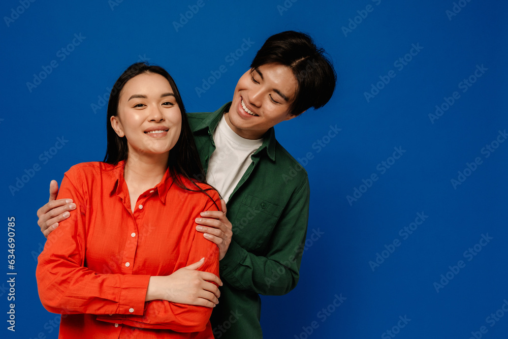 Smiling asian couple hugging while standing isolated over blue background