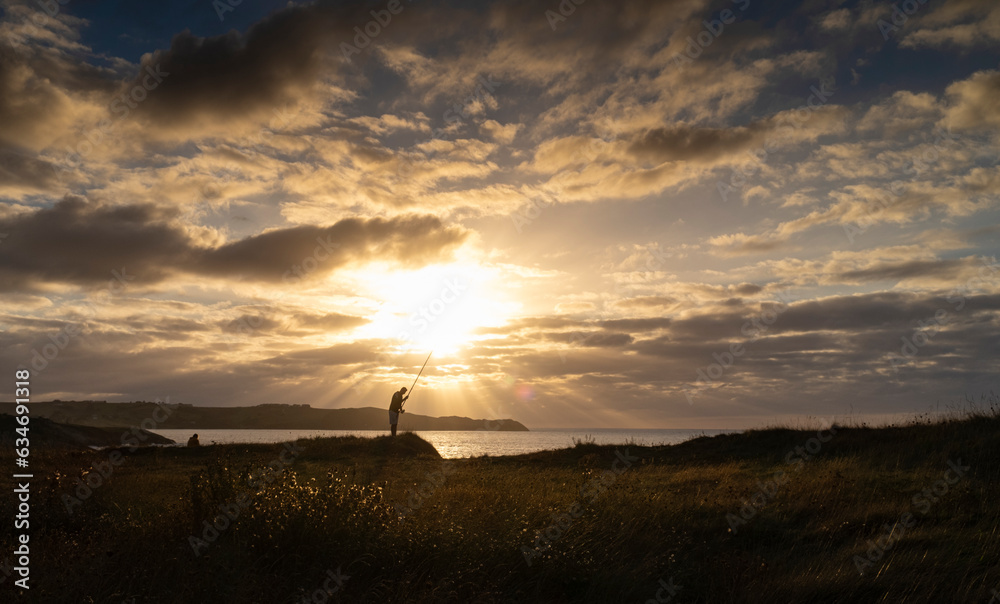 Fototapeta premium View of the coast at sunset over the Cantabrian Sea with the sun in front and the silhouette of a fisherman.