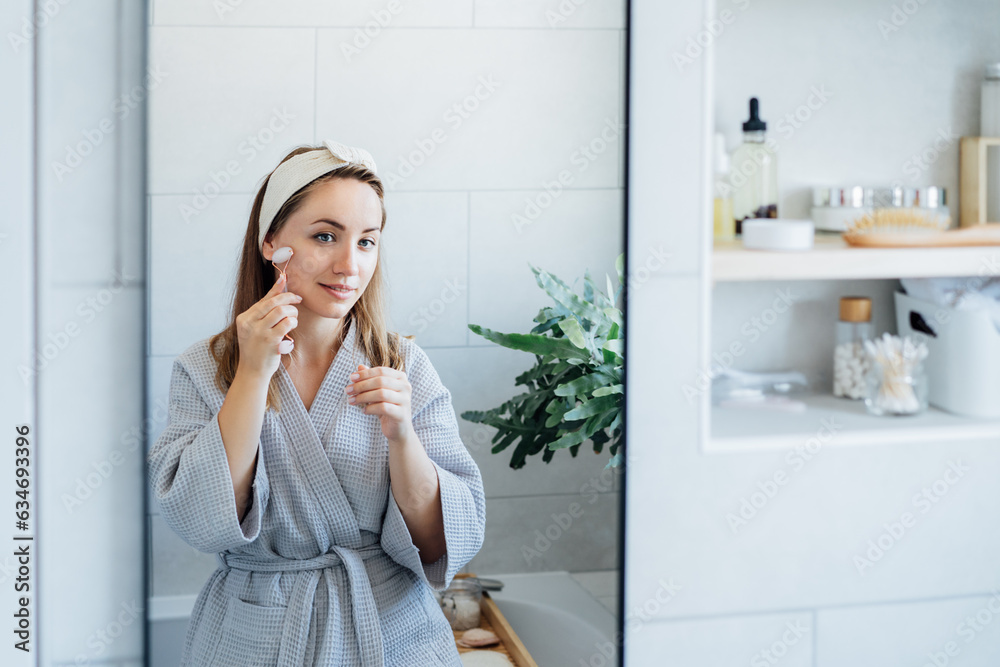 Young woman in bathrobe looking in mirror and making face massage with jade roller in the bathroom. The concept of natural procedures for skin care. Home Beauty self-care treatment. Selective focus Young woman in bathrobe looking in mirror and making face massage with jade roller in the bathroom. The concept of natural procedures for skin care. Home Beauty self-care treatment. Selective focus