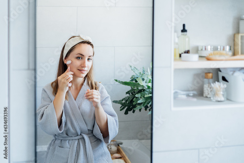 Young woman in bathrobe looking in mirror and making face massage with jade roller in the bathroom. The concept of natural procedures for skin care. Home Beauty self-care treatment. Selective focus