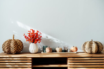 Cozy autumn home interior - various decorative wicker pumpkins, candles, seasonal flowers in vase on the wooden console with white wall background. Scandinavian minimalist hygge home fall decor.