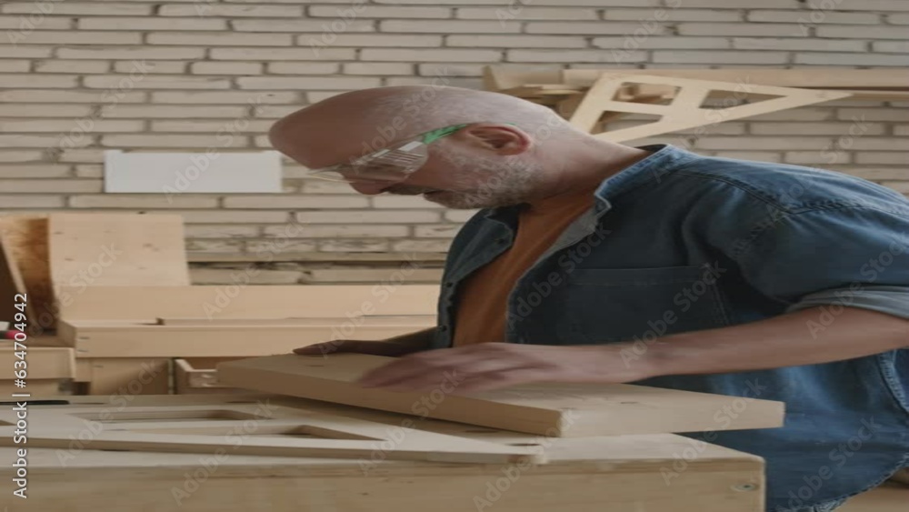 Vertical medium shot of adult Caucasian male carpenter measuring wooden plank before using wood cutter, making furniture in carpentry workshop Vertical medium shot of adult Caucasian male carpenter measuring wooden plank before using wood cutter, making furniture in carpentry workshop