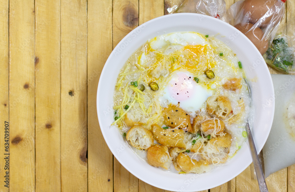 Hot congee or Rice porridge with soft boiled egg served in a white bowl with deep-fried dough stick (crispy patongo) on woodden backgrounds,left copy space fortext.