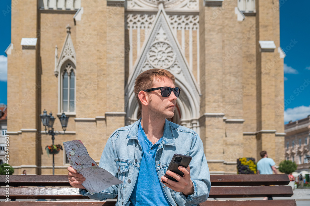 Man tourist sits on a bench in the city square with map and mobile phone and looks for hotel he booked from an application on his phone Male traveler 