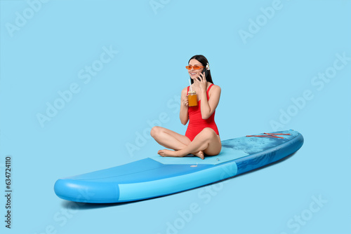 Happy woman with refreshing drink resting on SUP board and talking on smartphone against light blue background