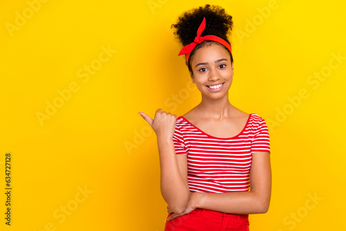 Photo of adorable cheerful girl dressed red striped t-shirt pointing thumb empty space isolated yellow color background