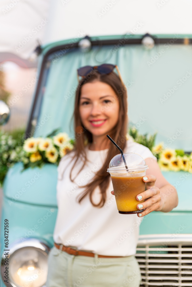 Young Happy woman holding a cold summer drink in front of food truck. Selective focus. Caucasian girl holding a cup of Bumble coffee to go at mint food truck. Street food business concept. Vertical Young Happy woman holding a cold summer drink in front of food truck. Selective focus. Caucasian girl holding a cup of Bumble coffee to go at mint food truck. Street food business concept. Vertical