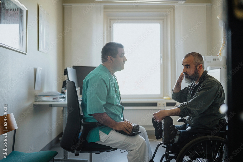 Side view of mature male doctor discussing with patient sitting in wheelchair at clinic Side view of mature male doctor discussing with patient sitting in wheelchair at clinic