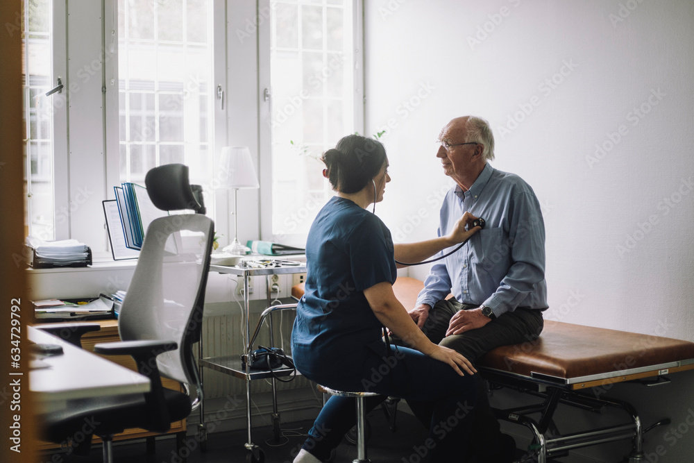 Female healthcare worker examining senior patient sitting on bed in clinic Female healthcare worker examining senior patient sitting on bed in clinic