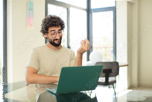young adult bearded man with a laptop making capice or money gesture, telling you to pay your debts!