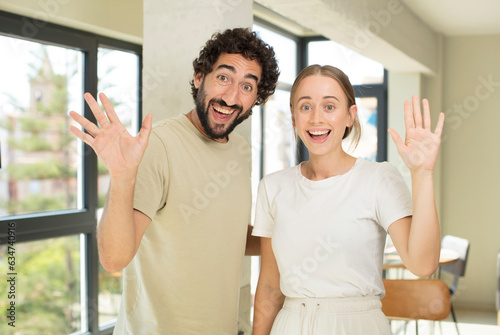 young adult couple smiling happily and cheerfully, waving hand, welcoming and greeting you, or saying goodbye