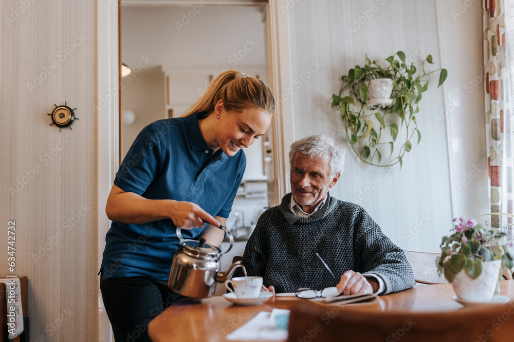 Smiling care assistant serving tea to senior man sitting at home Smiling care assistant serving tea to senior man sitting at home